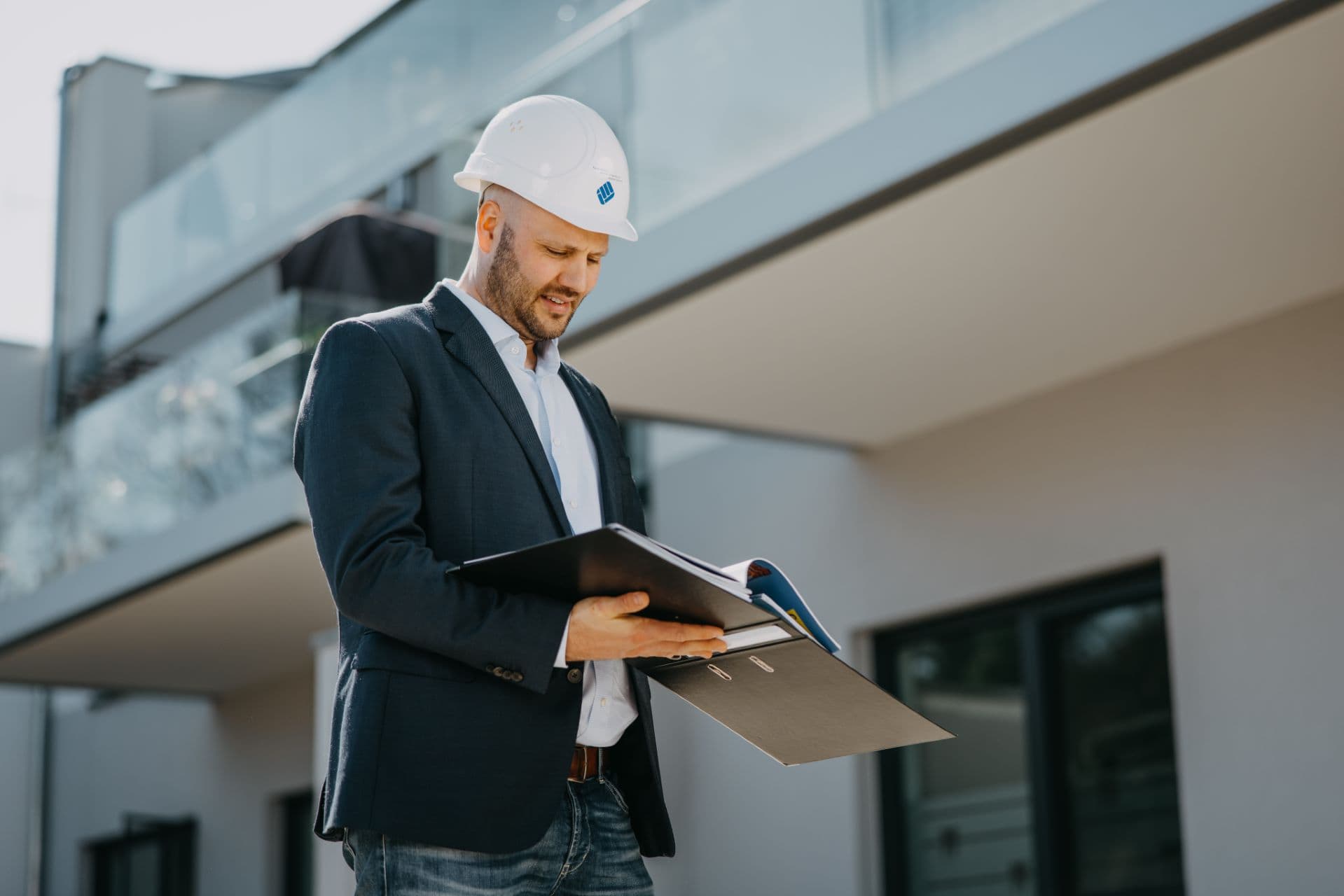 Baumeister mit Bauhelm prüft Baupläne vor einem modernen Neubau, Investment in nachhaltiges Bauprojekt, Architekturstil, Bauleitung, Bauvorhaben, Stadtentwicklung, Infrastruktur, Bauinspektion, Gebäudetechnik.