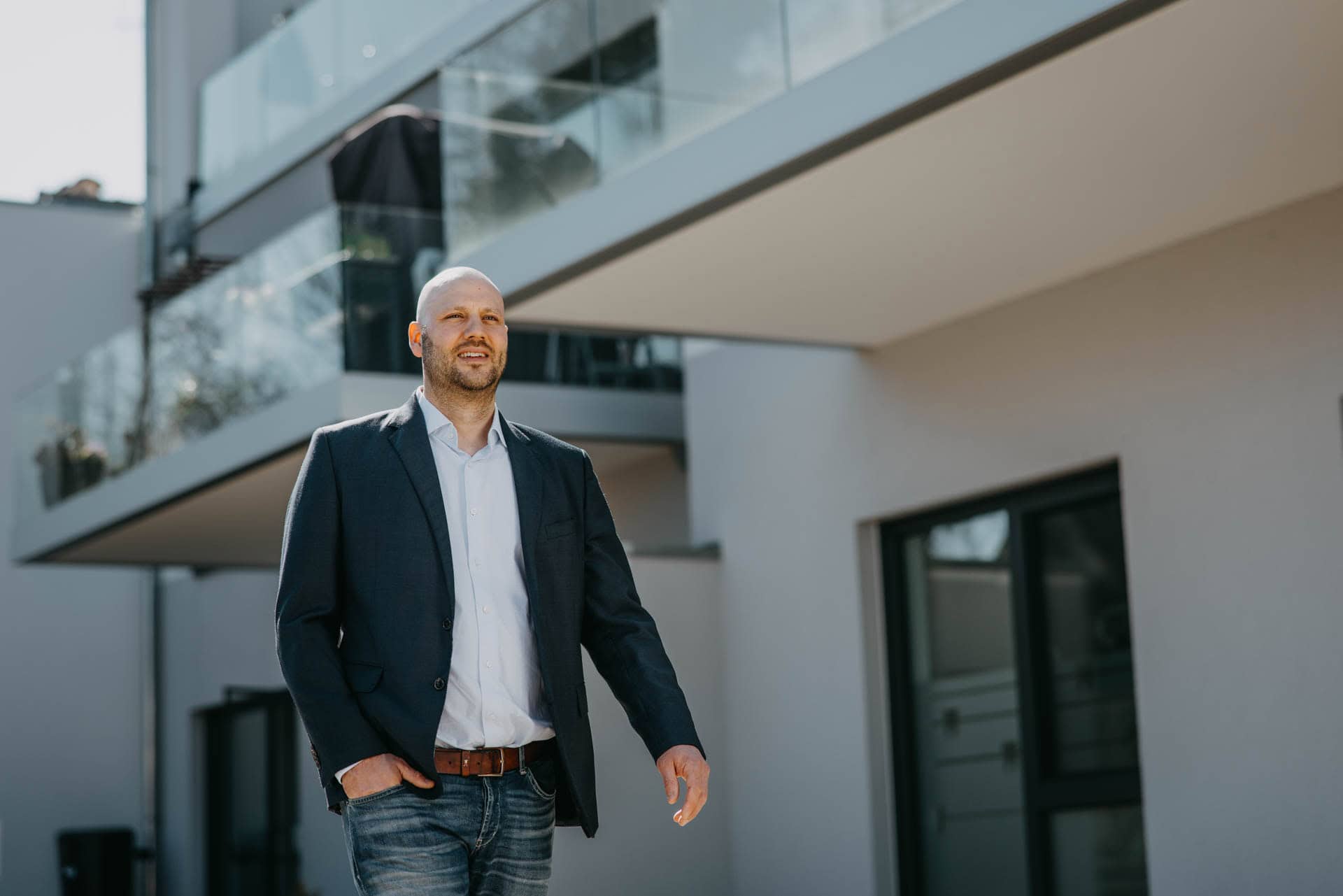 Modern gekleideter Mann in Anzug vor einem modernen, weißen Wohnhaus mit Balkon, Tageslicht und klarem Himmel, unterwegs nach draußen, stilvoll und professionell wirkend, Stadt- oder Vorort-Umfeld, Business- oder Immobilien-Kontext.